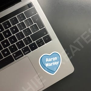 May include: A blue heart-shaped sticker with the text "Aaron Warner" on a silver laptop.