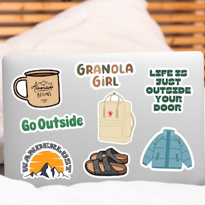 May include: A laptop with several stickers on it. The stickers feature a mug, a backpack, a jacket, and text that says "Granola Girl", "Go Outside", "Wanderlust", and "Life is just outside your door".