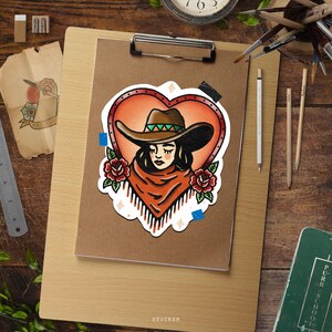 May include: A sticker featuring a cowgirl design within a heart-shaped frame. The cowgirl wears a brown hat with a green band, a fringed orange shawl, and is surrounded by red roses. The word "STICKER" is at the bottom.