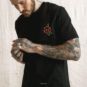 May include: Black t-shirt with a red rose and green leaves embroidered on the chest. The word "TSHIRT" is printed at the bottom of the image. The shirt is made of a soft material.