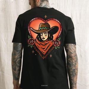 May include: Black t-shirt featuring a tattoo-style design on the back. The design includes a cowgirl wearing a brown hat and a red scarf, set within a heart-shaped frame. Red roses and stars surround the central image. The word "TSHIRT" is printed at the bottom.