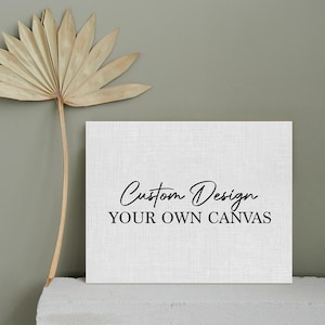 May include: A white canvas with the text "Custom Design YOUR OWN CANVAS" in black script. A dried palm leaf is positioned to the left of the canvas. The canvas is on a white surface against a gray-green background.