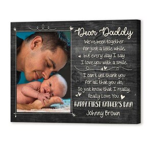Puede incluir: Impresi&oacute;n en lienzo enmarcada con una foto de un padre y un beb&eacute;, con el texto "Dear Daddy" y un mensaje del D&iacute;a del Padre. El fondo es un patr&oacute;n de vetas de madera oscura.