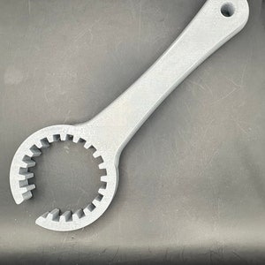 May include: A light gray, 3D-printed spanner wrench. The wrench has a long handle with a hole at the end for hanging. The head of the wrench has a circular opening with teeth around the inside edge. The tool is designed for tightening or loosening nuts and bolts.