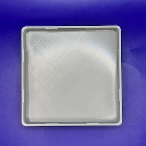 May include: A light gray, square tray with a textured surface and a raised border. The tray has a simple, minimalist design and is likely made of plastic or a similar material. The corners are rounded, and the edges have small cutouts.
