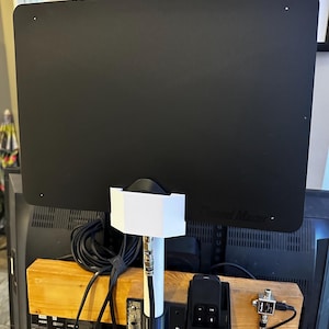 May include: A black Channel Master TV aerial is mounted on a wooden stand. The aerial is rectangular with rounded corners. Cables and a black remote control holder are attached to the stand. A world map is visible in the background.