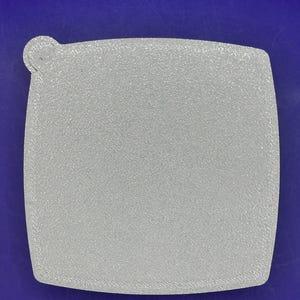 Puede incluir: Un objeto cuadrado gris claro con esquinas redondeadas y una pequeña extensión circular en un lado. El objeto tiene una superficie texturizada. El fondo es de color azul liso.