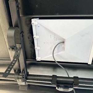 Può includere: Un dispositivo bianco di forma geometrica è montato su un telaio metallico nero. Un cavo nero si estende dal dispositivo. Il dispositivo è fissato con cinghie nere e staffe metalliche. L'immagine è ripresa dal basso.