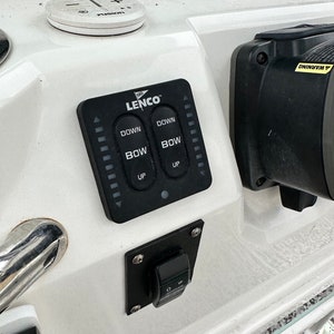 May include: A boat control panel featuring a black Lenco trim tab switch with "DOWN" and "BOW" buttons. A small, round Fusion audio control is also visible. The panel is set on a white surface, typical of marine equipment.