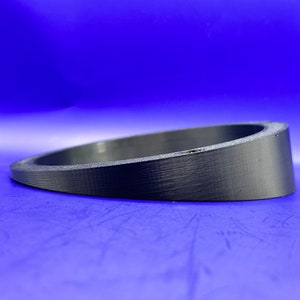 May include: A black, circular, angled object with a textured surface. The object is set against a vibrant blue background. The shape suggests a ring or a spacer, possibly for mechanical or decorative purposes.