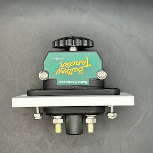 Könnte beinhalten: Ein schwarzes und grünes Battery Tender-Gerät mit einer weißen Montageplatte. Das Gerät hat einen schwarzen Knopf, und der Text "Battery Tender" ist auf der grünen Fläche sichtbar. Die Website BatteryTender.com ist ebenfalls sichtbar.