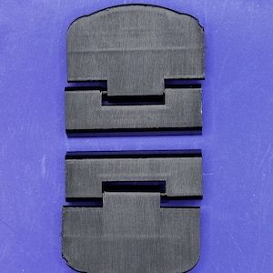 Puede incluir: Dos objetos rectangulares negros y texturizados con una superficie acanalada, dispuestos sobre un fondo azul. Cada objeto tiene una forma única con una base rectangular y una parte superior curvada. Los objetos parecen estar hechos de un material duradero.