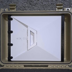 Può includere: Una valigia rettangolare rigida color beige con un telaio interno nero. All'interno, è visibile una struttura bianca angolata con un ritaglio rettangolare. La valigia è aperta, rivelando il contenuto.