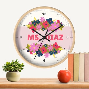 Puede incluir: Un reloj de pared redondo con marco de madera clara. La esfera es blanca con números y manecillas negras. El centro presenta el texto "MS. DIAZ" en rosa, rodeado de arreglos florales.