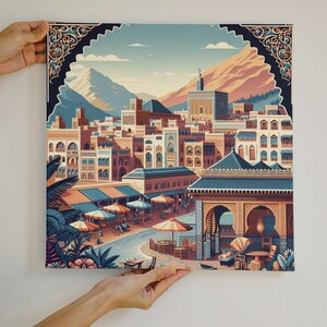 Puede incluir: Impresión en lienzo cuadrada que representa una ciudad estilizada con edificios, montañas y cielo azul. La obra de arte utiliza azules, naranjas y marrones, con arcos y toldos decorativos. La impresión está sujeta por dos manos.