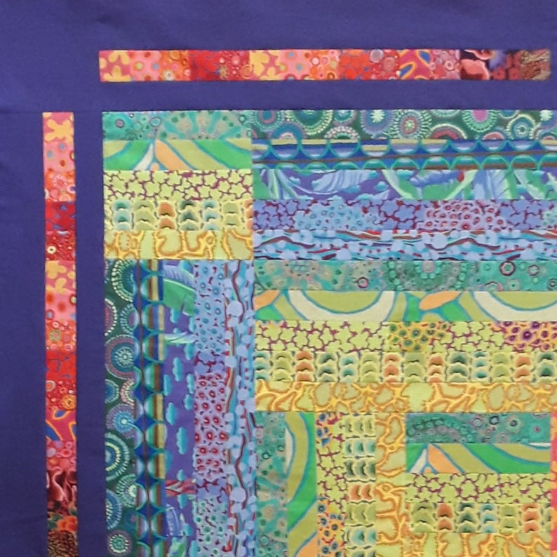 May include: A purple fabric background with a colorful patchwork quilt design. The quilt is made of various fabrics with different patterns, including circles, dots, and stripes. The quilt is bordered with a thin red and orange strip.