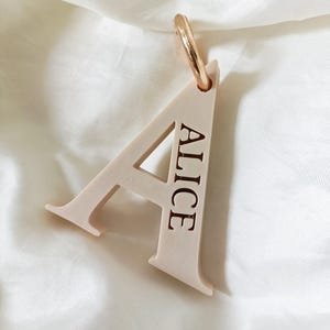 May include: A pale beige letter A pendant featuring the name ALICE engraved in a dark font along the right side of the letter. The pendant includes a small, circular, metallic rose gold jump ring attached to the top for hanging purposes.