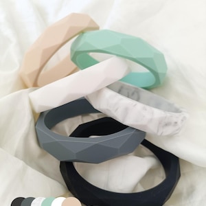 May include: A selection of geometric silicone bracelets in colours such as beige, mint green, white, marble, grey, and black. The bracelets have a faceted design and are stacked on a white cloth.