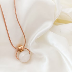 May include: A white stone pendant necklace with a gold-toned metal clasp and a brown cord. The necklace is displayed on a white satin fabric.