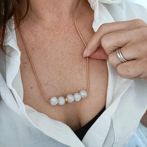 A gold cord chain necklace with a row of five white silicone pearls and a gold accent piece.