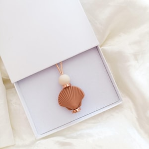 Könnte beinhalten: Eine braune Muschel-Anhänger-Kette mit einer weißen Perle und einer roségoldenen Kette in einer weißen Schachtel.