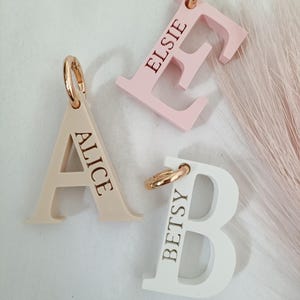 May include: Three letter-shaped bag charms with rose gold-toned metal rings. One beige letter A engraved with ALICE, one light pink letter E engraved with ELSIE, and one white letter B engraved with BETSY. Each charm features a smooth finish.