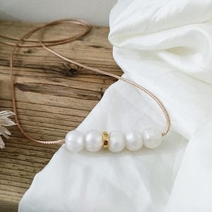 May include: A simple necklace with a gold accent and five white pearls. The necklace is on a light brown wooden surface with a white fabric in the background.