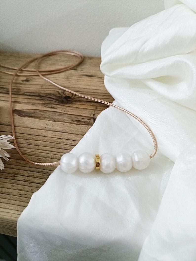May include: A simple necklace with a gold accent and five white pearls. The necklace is on a light brown wooden surface with a white fabric in the background.