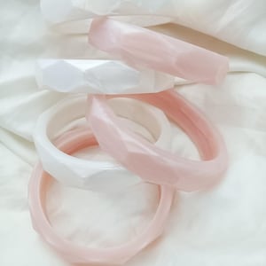 May include: A collection of white and pink geometric bangles. The bracelets are stacked on a white, textured surface. The bangles have a faceted design, and the pink ones have a translucent appearance.