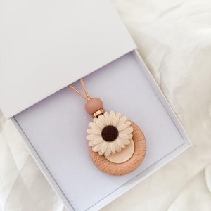 Könnte beinhalten: Eine Holz-Anhänger-Halskette mit Gänseblümchen-Design, präsentiert in einer weißen Box. Die Halskette hat einen hellbraunen Holzring, eine cremefarbene Gänseblümchen und eine hellbraune Kordel. Das Gänseblümchen hat eine dunkelbraune Mitte.