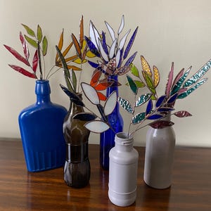 Può includere: Una collezione di steli di foglie decorative in vetro colorato, esposte in bottiglie di diversi colori. Le foglie sono rosse, verdi, arancioni, gialle, bianche e blu. Le bottiglie sono blu, marroni, bianche e grigie.