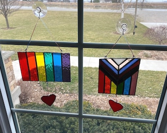 May include: Two stained glass window hangings with red hearts. The first is a rainbow flag design. The second is a geometric design with a rainbow color scheme.