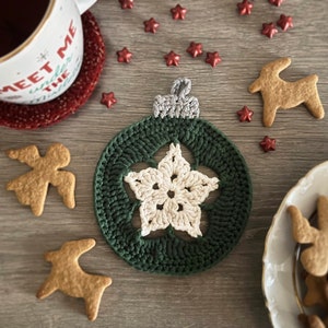May include: A green crocheted Christmas ornament with a white star in the center. The ornament is on a wooden surface with gingerbread cookies and red glitter stars.