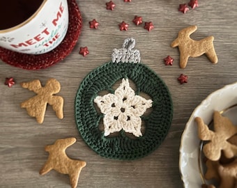 Puede incluir: Un adorno navideño de crochet verde con una estrella blanca en el centro. El adorno está sobre una superficie de madera con galletas de jengibre y estrellas de purpurina roja.