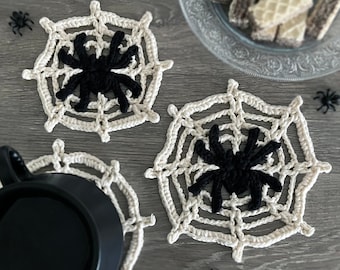 May include: Three crocheted coasters in the shape of spiderwebs with black spiders in the center. The coasters are on a gray wooden surface.