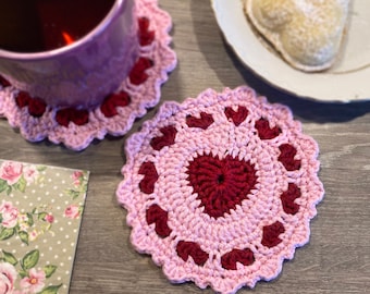 May include: Two pink and burgundy crocheted coasters with a heart design. The coasters are on a wooden surface.