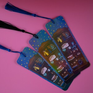 May include: Three illustrated bookmarks with blue tassels against a pink background. The bookmarks feature a night sky design with stars and a whimsical illustration of a cake, a creature, and other objects. One tassel is black.