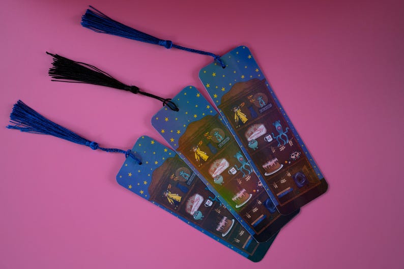 May include: Three illustrated bookmarks with blue tassels against a pink background. The bookmarks feature a night sky design with stars and a whimsical illustration of a cake, a creature, and other objects. One tassel is black.