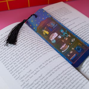May include: A colorful bookmark with a black tassel, placed in an open book. The bookmark features a blue background with stars and various cartoon illustrations. The book is open to a page with printed text.
