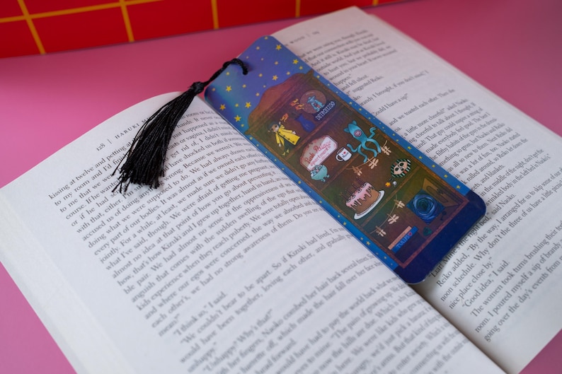 May include: A colorful bookmark with a black tassel, placed in an open book. The bookmark features a blue background with stars and various cartoon illustrations. The book is open to a page with printed text.