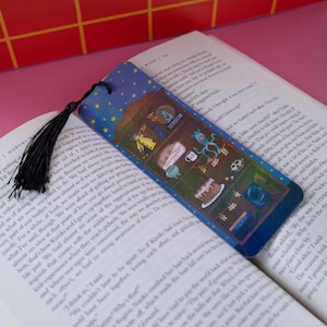 May include: A blue bookmark with a starry sky design and a black tassel. The bookmark features cartoon illustrations of various objects, including a cake, a cup, and a creature, and the word "DETROITEDO".