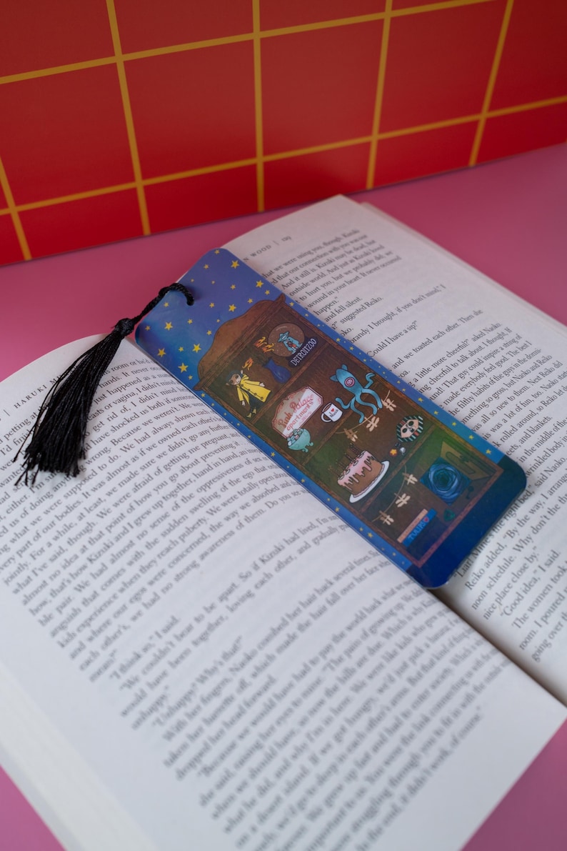 May include: A blue bookmark with a starry sky design and a black tassel. The bookmark features cartoon illustrations of various objects, including a cake, a cup, and a creature, and the word "DETROITEDO".