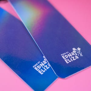 May include: Two blue rectangular tags with iridescent color gradients are displayed against a pink background. The tags have rounded corners and a glossy finish. The text "MADE BY EDNA & ELIZA" is printed on each tag in white.