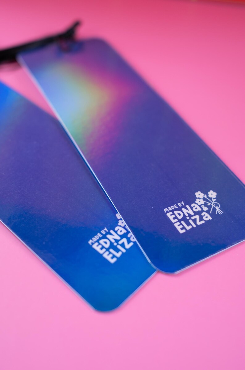 May include: Two blue rectangular tags with iridescent color gradients are displayed against a pink background. The tags have rounded corners and a glossy finish. The text "MADE BY EDNA & ELIZA" is printed on each tag in white.