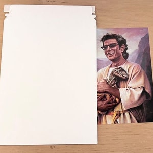 May include: A print of a man wearing glasses and a light-colored robe, holding a small dinosaur. The background features a mountain range and a purple sky. The print is partially visible next to a white envelope.