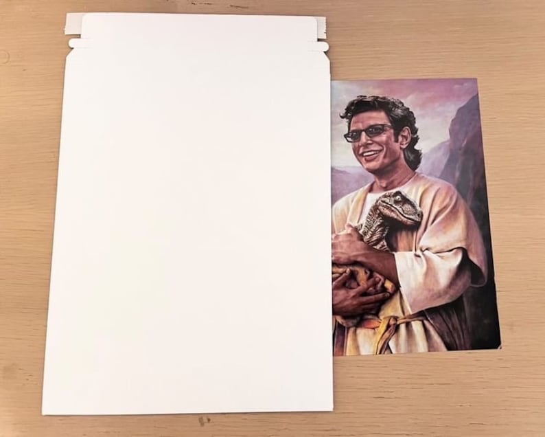 May include: A print of a man wearing glasses and a light-colored robe, holding a small dinosaur. The background features a mountain range and a purple sky. The print is partially visible next to a white envelope.