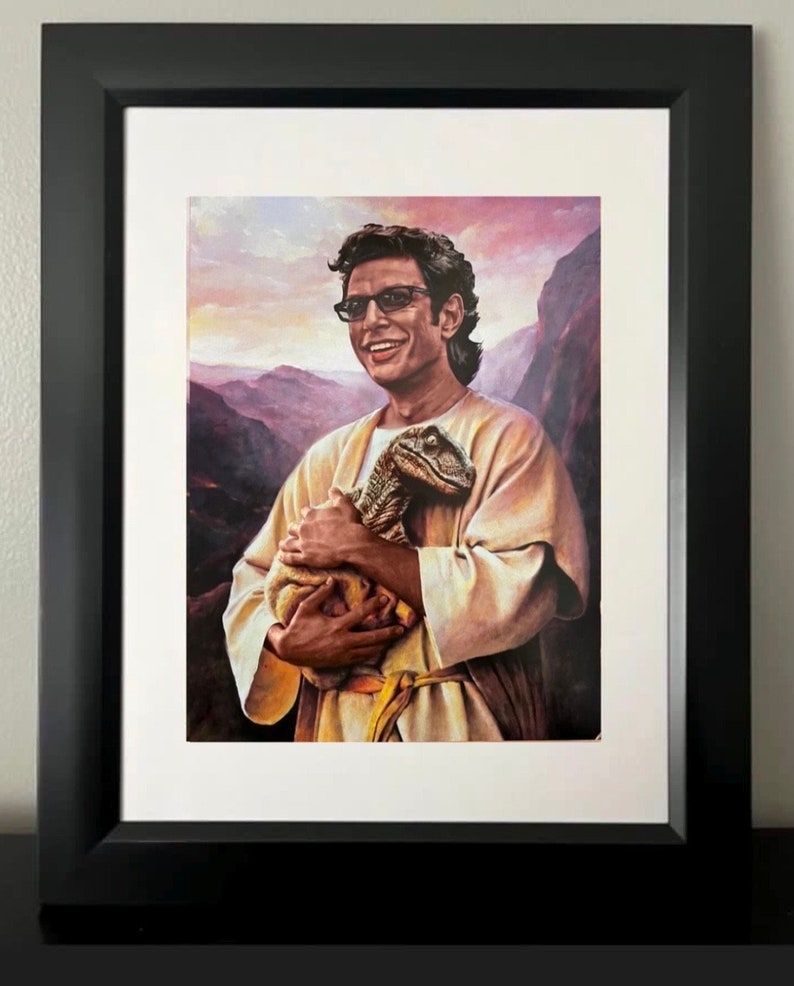 May include: Framed print featuring a portrait of a man holding a dinosaur. The man is wearing glasses and a light-colored robe, set against a mountainous backdrop. The print is in a black frame with a white mat.