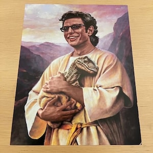 May include: A print featuring a man in a light-colored robe holding a baby dinosaur. The man is wearing glasses and smiling. The background shows a mountain range and a sunset. The overall theme is a humorous take on a religious painting.