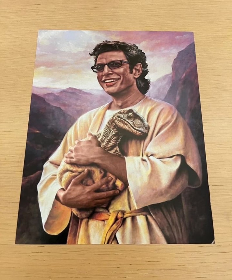 May include: A print featuring a man in a light-colored robe holding a baby dinosaur. The man is wearing glasses and smiling. The background shows a mountain range and a sunset. The overall theme is a humorous take on a religious painting.