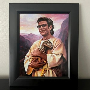 May include: Framed print featuring a portrait of a man in sunglasses holding a small dinosaur. The artwork is set against a backdrop of a mountain landscape with a sunset. The frame is black.